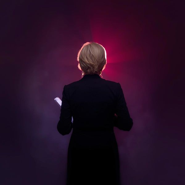 A female actor about 50 years old reading from a script wearing all black with a coloured light on them from the waist up from behind